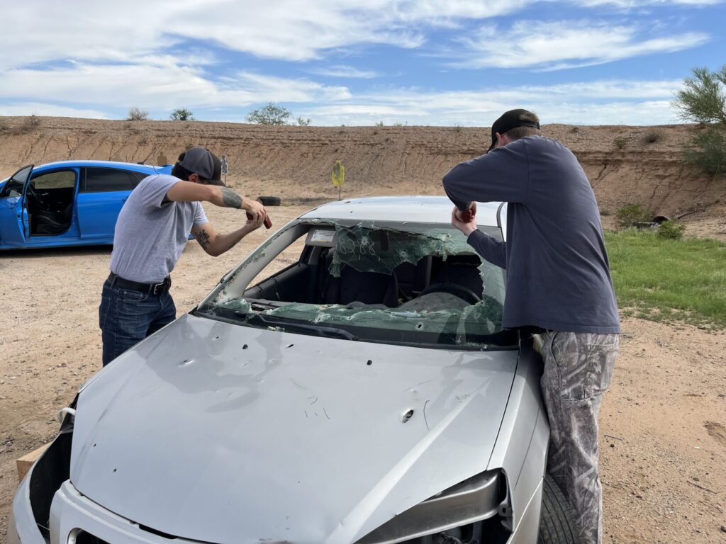 Image of two men leaning over the hood of a shot-up car on a firing range. They are standing beside the front fenders, just ahead of the front seat doors. They are holding their pistols over the front seats, pointed down at the "laps" of those who would be sitting in the seats. They are holding their support hand palms in front of, rather than beside, the grips of their pistols.