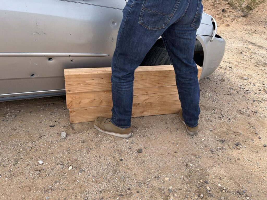 Image of a man's legs in bluejeans. He is protected from ricochets by three stout 4x4s, stacked one upon the other.