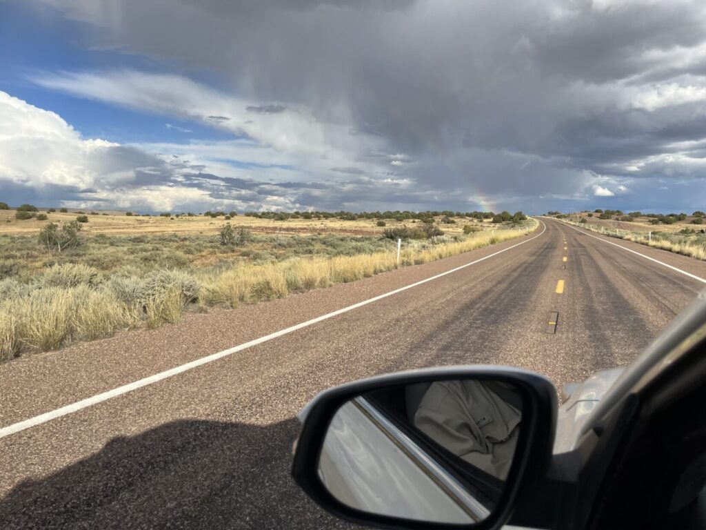 Image from a driver's side car window. the road winds up and to the left across reset grasslands. Scattered clouds and a rainbow are overhead.
