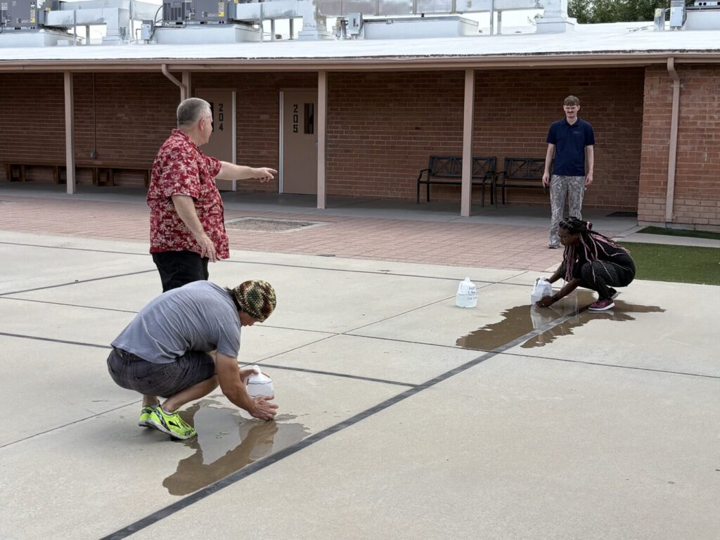 Two Bleeding Control students apply pressure to jugs in a school courtyard, while two instructors look on.