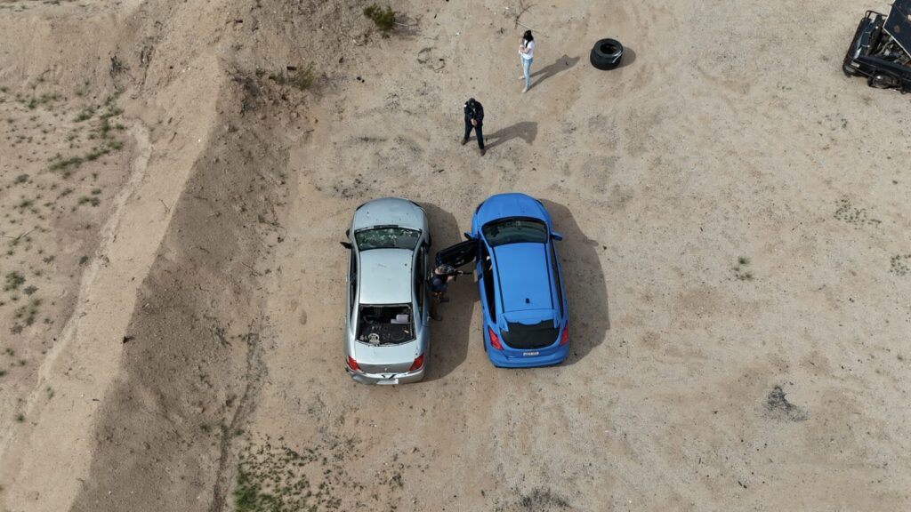 Image from above and behind a pair of parked sedans. The car on the right is blue. The driver's door of the blue car is open. Two re-enactors, playing A T F agents, 90 degrees offset from one another, point plastic pistol replicas at the car. A woman holding an infant CPR manikin stands off the the side role playing the victim of a carjacking.
