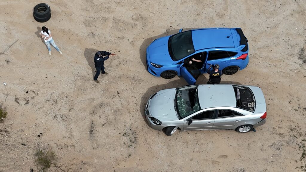 Image of two cars parked side by side as viewed from above. The blue sedan on the right has the driver's side door open. A role player in police raid gear points an H&K MP5 submachine gun (with a barrel insert for safety) at the driver through the open door. Another role player points a (plastic replica) pistol through the windshield from a few feet in front of the car. A woman playing the recently carjacked mother stands off to the side, holding an infant CPR manikin.