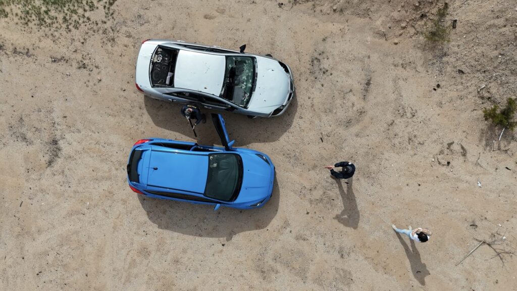 Image of two cars parked side by side as viewed from above. The blue sedan on the right has the driver's side door open. A role player in police raid gear points an H&K MP5 submachine gun (with a barrel insert for safety) at the driver through the open door. Another role player points a (plastic replica) pistol through the windshield from a few feet in front of the car. A woman playing the recently carjacked mother stands off to the side, holding an infant CPR manikin.
