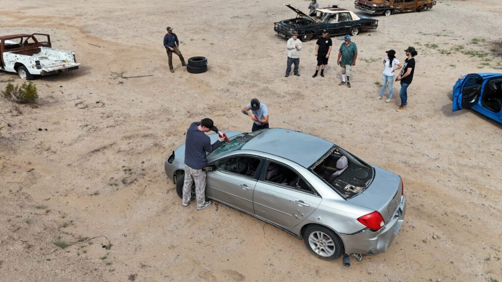 Aerial photo of two shooters demonstrating where to stand, just ahead of the rearview mirrors of a silver sedan. Several observers watch.