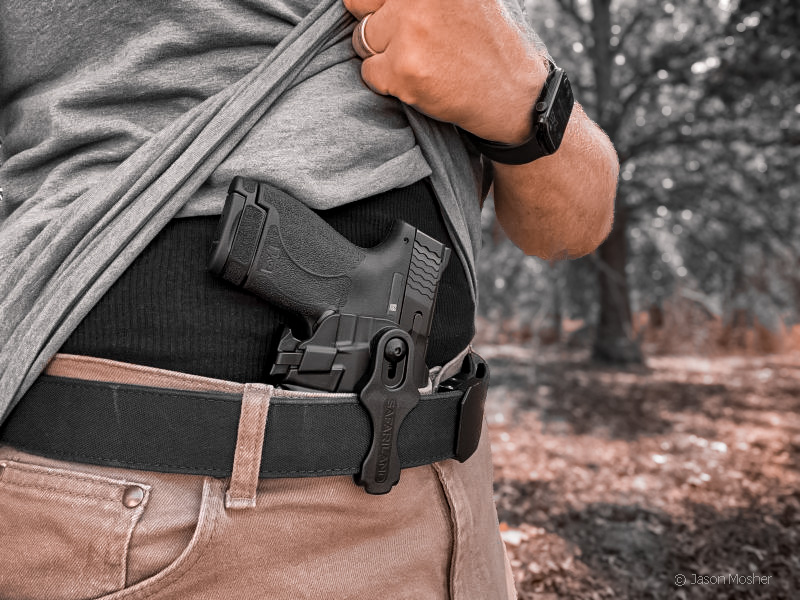A person in a grey shirt, pulling the shirt out of the way to expose a black handgun in a 575 Pro-Fit GLS IWB holster attached to belt around waist.