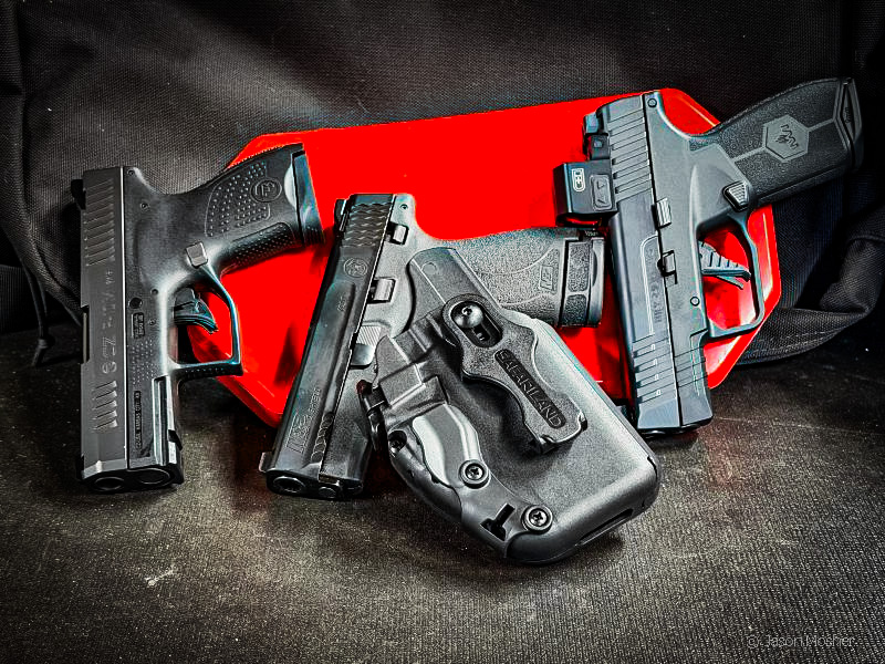 Multiple, compact black handguns laying against a red surface with a Pro-Fit holster in front of them.
