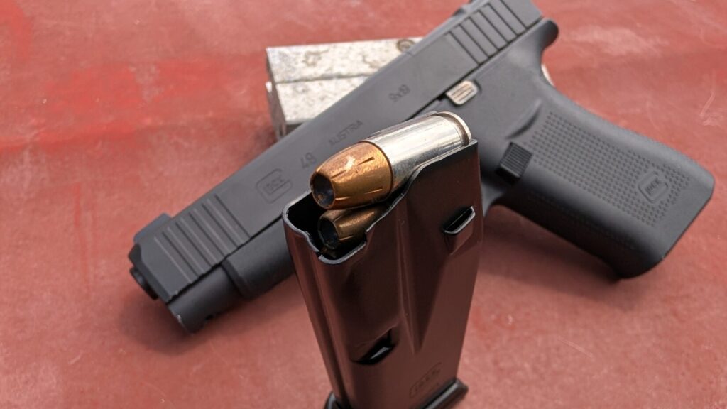 Loaded Glock magazine standing upright in front of a Glock 48 pistol against a wooden backdrop.