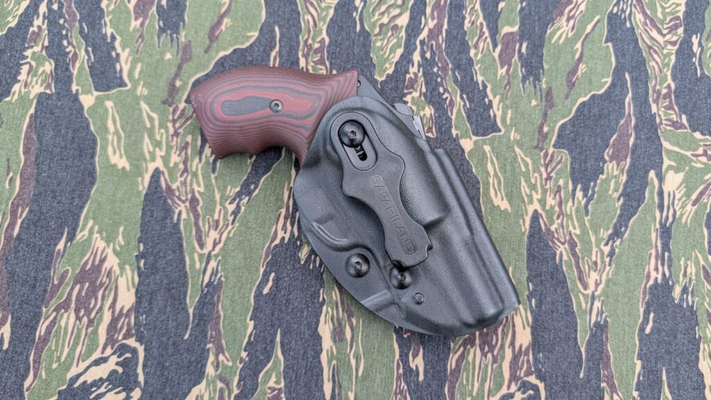 Angled view of the front of a black Safariland holster with clip attached, resting on camo fabric.