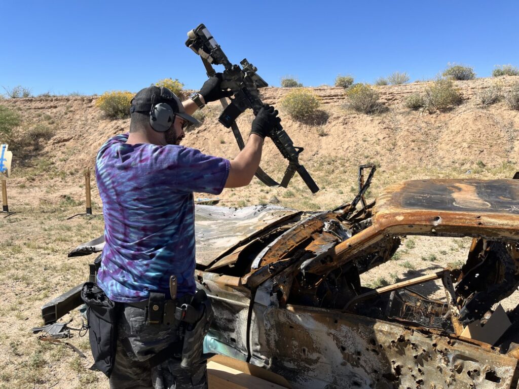 Image of a man in a blue and purple tie-dye shirt holding a suppressed A-R series rifle over the windshield area of a shot-up junk car.