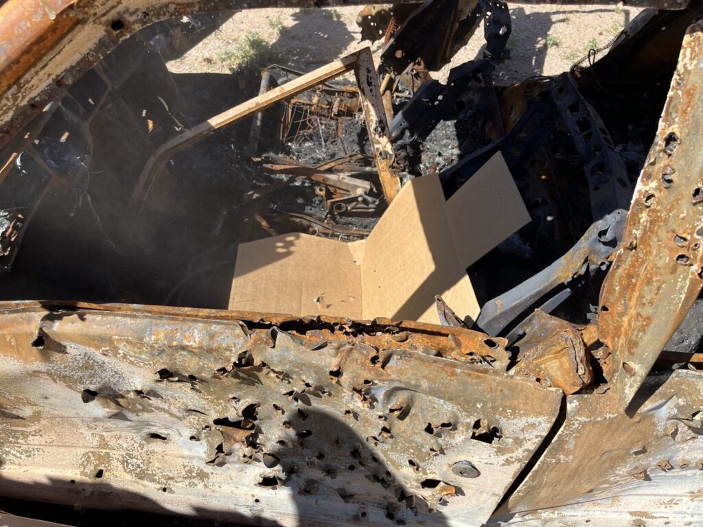 Image of a shot up junk car with a cardboard silhouette target in the driver's seat, folded forward at the bottom as if sitting.