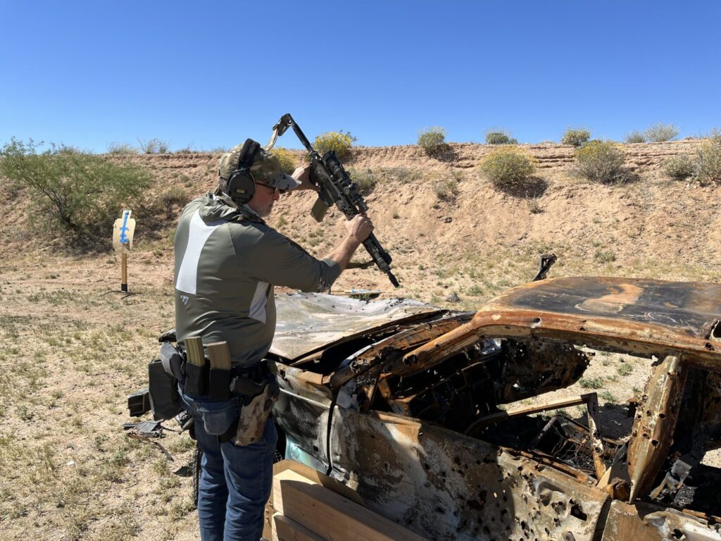Image of a shooter standing by the front left fender of a shot up junk car, pointing an A-R series rifle at the driver's seat. He has the thumb of his left hand against the side of the magazine well in front of the trigger area for safety.