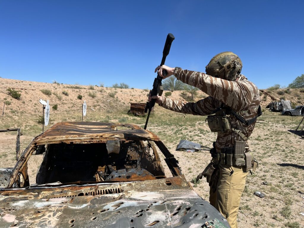 Image of a man in battle gear standing next to the front left fender of a shot up junk car, holding a rifle over the car pointed down into the bottom of the driver's seat.