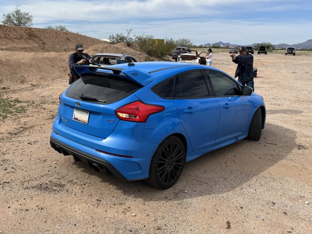 Image of a blue car. Role players acting as police officers stand beside and in front of the car, leveling replica pistols at it.
