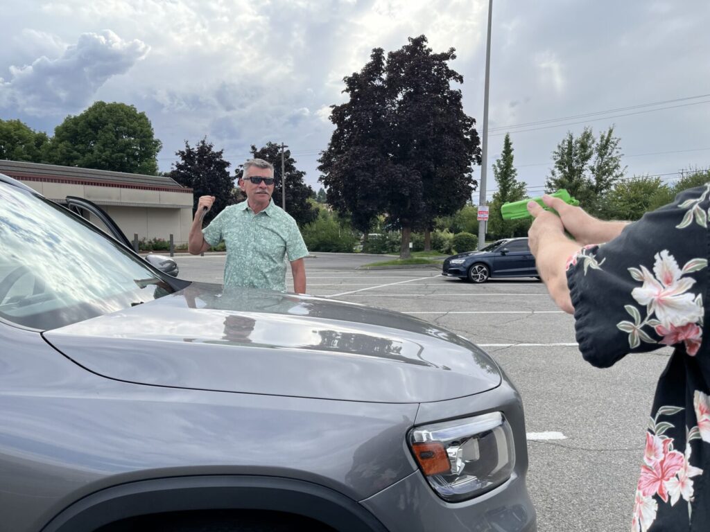 A man role-playing a carjacker stands on the opposite side of a car, brandishing a (rubber) knife. An armed citizen role player points a green plastic pistol replica at him over the hood.