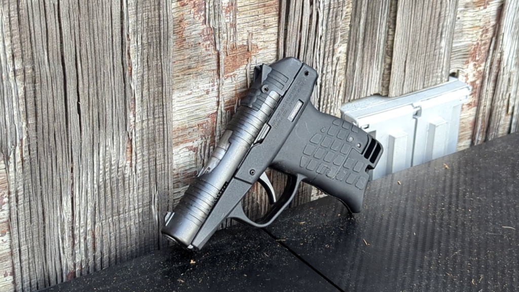 Right-side view of a black PR-3AT pistol propped upright against a rustic weathered wood plank wall.
