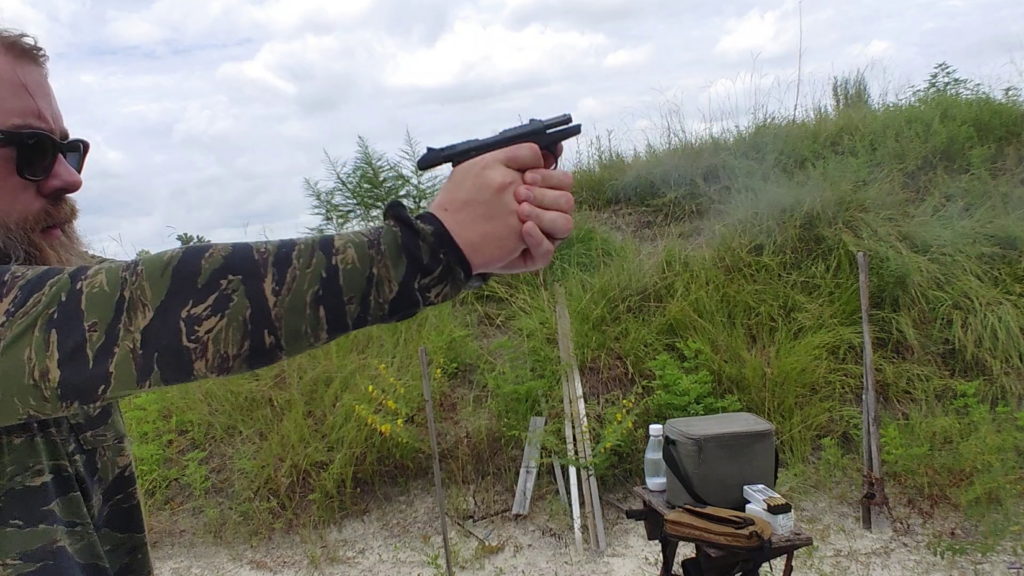 A person in a tiger-stripe camo jacket firing a black PR-3AT pistol with the slide locked back at an outdoor range.