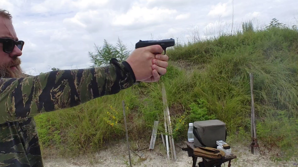 Side view of a shooter aiming a compact black PR-3AT pistol downrange against a grassy hill.