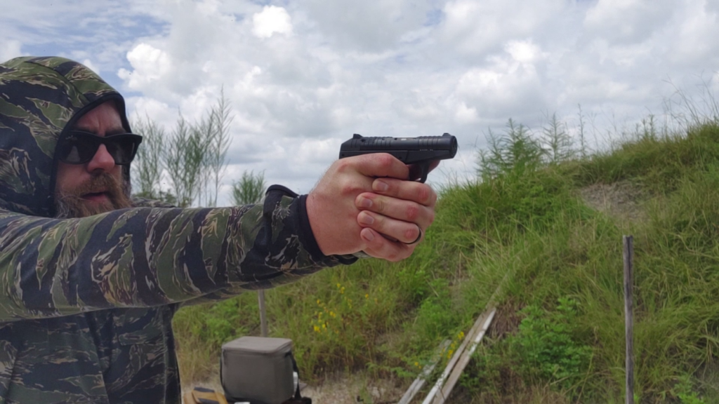 Shooter wearing sunglasses and a tiger-stripe camo hoodie gripping and aiming a black PR-3AT pistol forward.