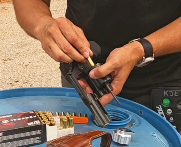 A man in a black shirt standing in front of a barrel loads a revolver one cartridge at a time.