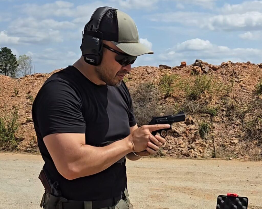A man in a black shirt holds a revolver at his centerline at a shooting range.