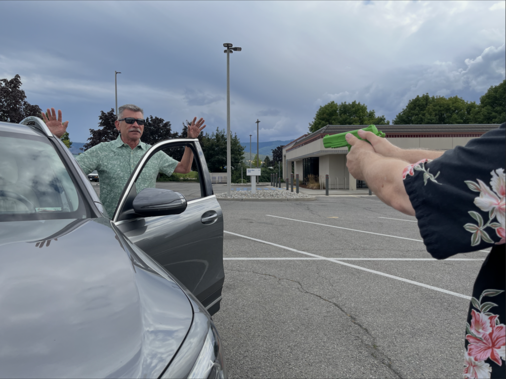 Image of a carjacker stepping out of car with his hands up. The car's owner keeps him covered while backing up to create distance. In this still photo, the armed citizen is about parallel with the front bumper.