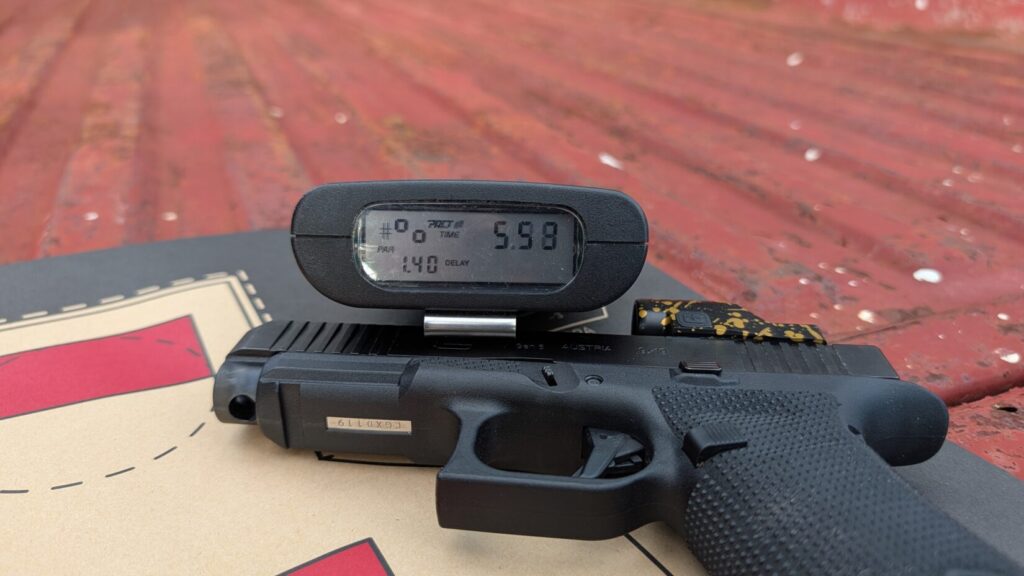 A close-up of a black shot timer resting on top of a black Glock handgun equipped with a red dot sight, laid on a paper target.
