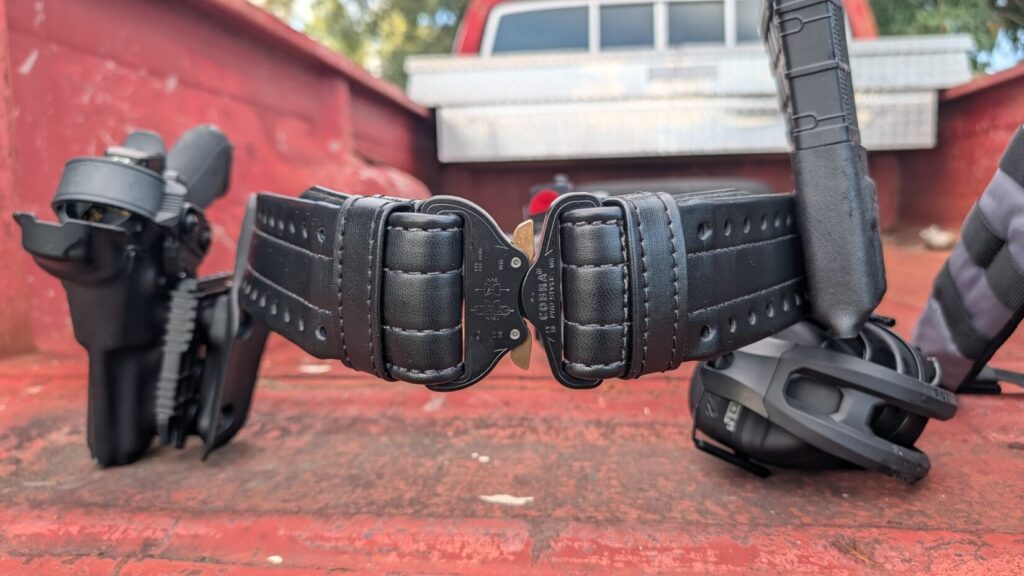 close-up of a black HOLLE belt fastened with a black Cobra buckle, with a holster, magazine pouch, and hearing protection nearby.