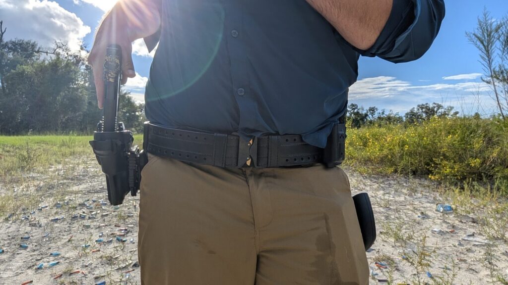 A person wearing a blue shirt and khaki pants outdoors, drawing a handgun from a black holster attached to a black HOLLE belt featuring a Cobra buckle.