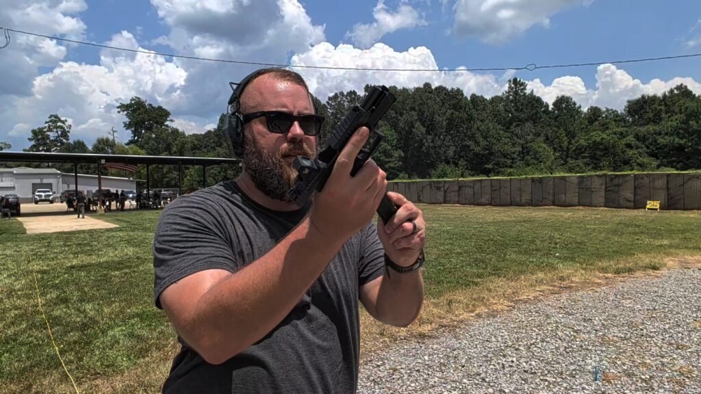 Travis Pike wearing sunglasses and ear protection reloading a CZ Duty at an outdoor shooting range.