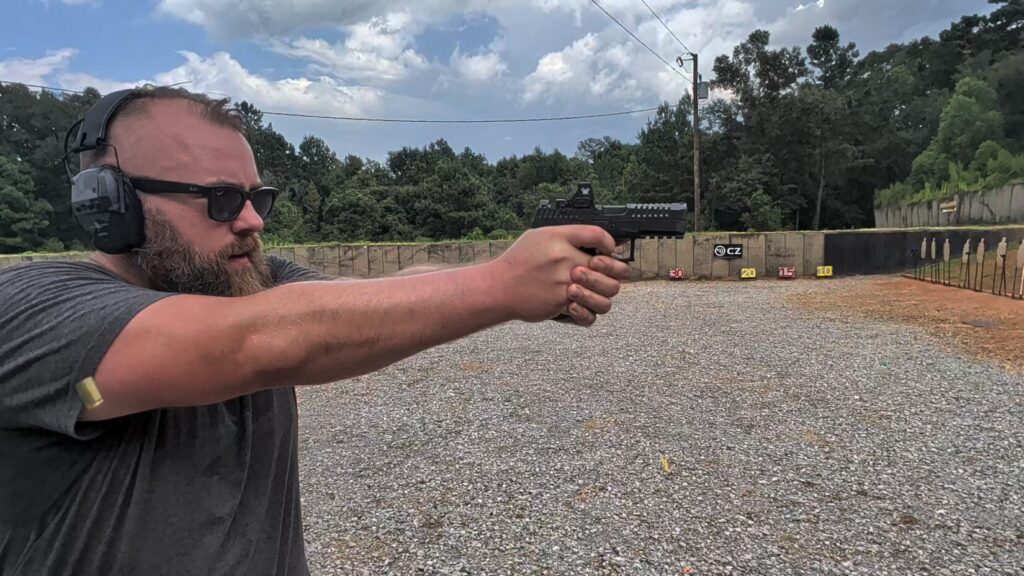 Travis Pike firing a CZ DUTY pistol at an outdoor range as a spent casing ejects past his arm.