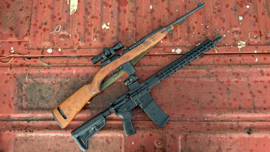 A scoped M1 Carbine rifle with a wooden stock lying next to a modern black AR-15-style rifle on a rusted red metal surface.