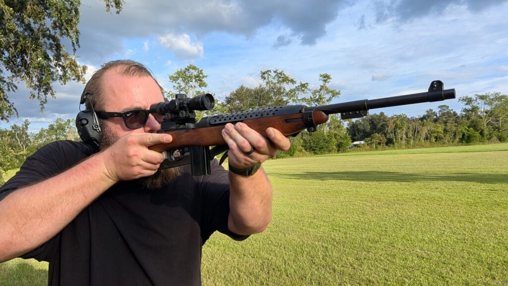 A man wearing sunglasses and ear protection standing and aiming a scoped M1 Carbine rifle outdoors in a green, grassy field.