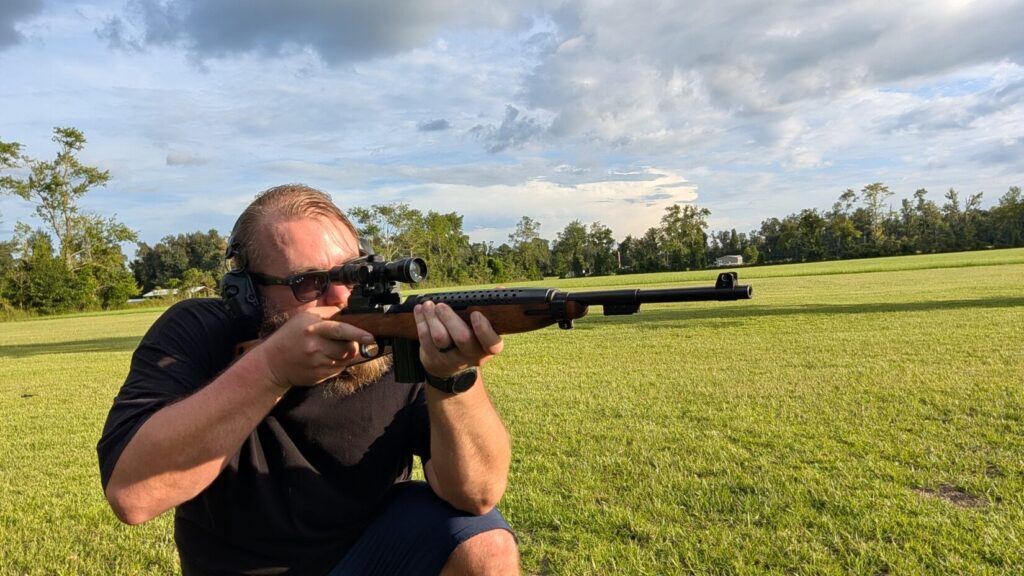 A man wearing sunglasses and ear protection kneeling while aiming a scoped M1 Carbine rifle outdoors in a grassy field.