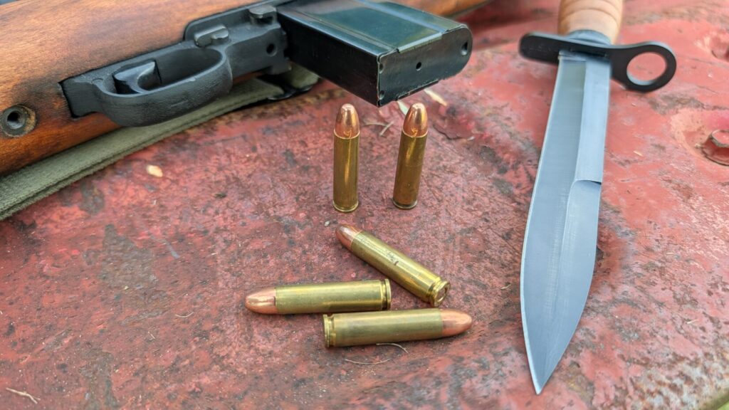 A detailed close-up of the receiver, trigger assembly, magazine, and scope of an M1 Carbine rifle resting on a rusty red metal surface.