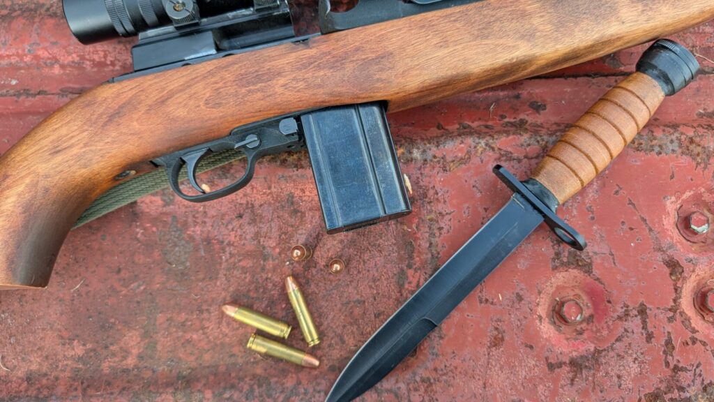 A close-up of an M1 Carbine rifle's receiver and magazine, three loose brass cartridges, and a black blade knife with a stacked leather handle on a rusty red surface.