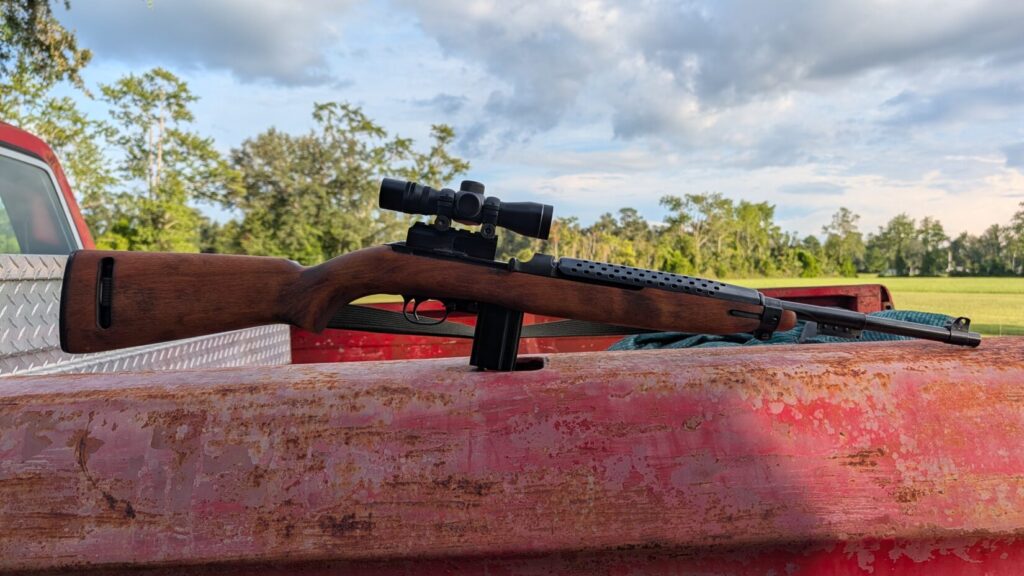 A scoped M1 Carbine rifle resting on the edge of a rusty red truck bed, with a green field, trees, and a cloudy sky in the background.