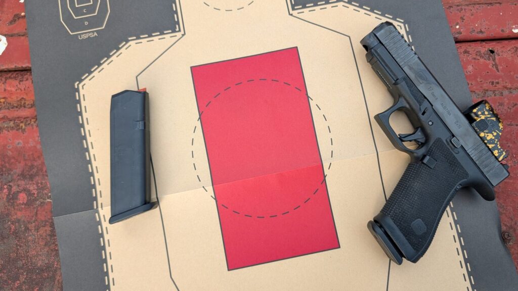 A black Glock pistol equipped with a red dot sight and a spare magazine lying on a cardboard target with a red rectangular center.