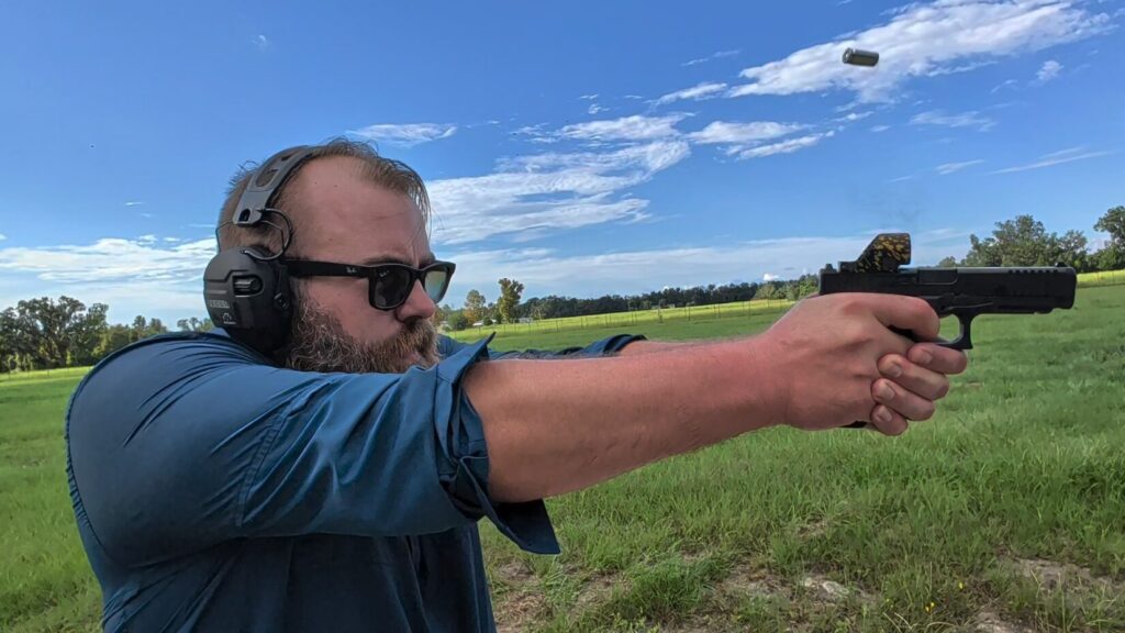 Side profile of a bearded man wearing ear and eye protection firing a handgun outdoors, with an ejected spent casing visible in mid-air.