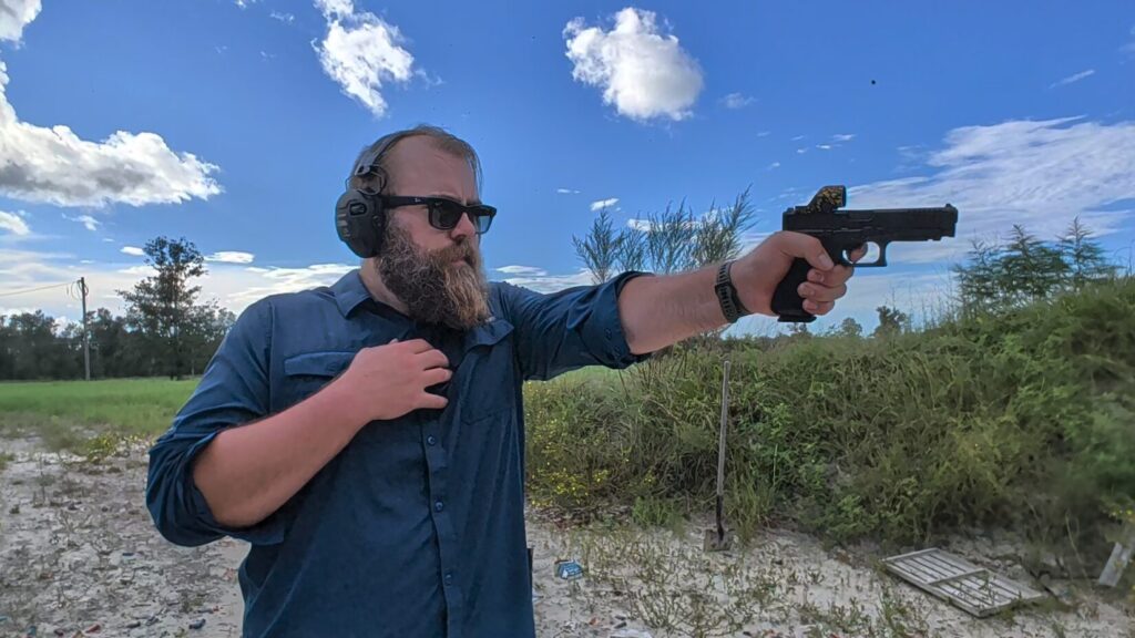 Close-up of a bearded man in safety gear aiming a handgun with his right hand while holding his left hand tightly against his chest.