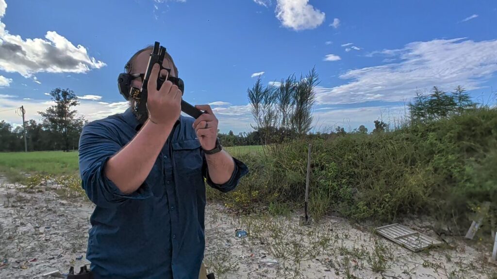 A man wearing ear and eye protection reloads a black semi-automatic pistol at an outdoor shooting range.
