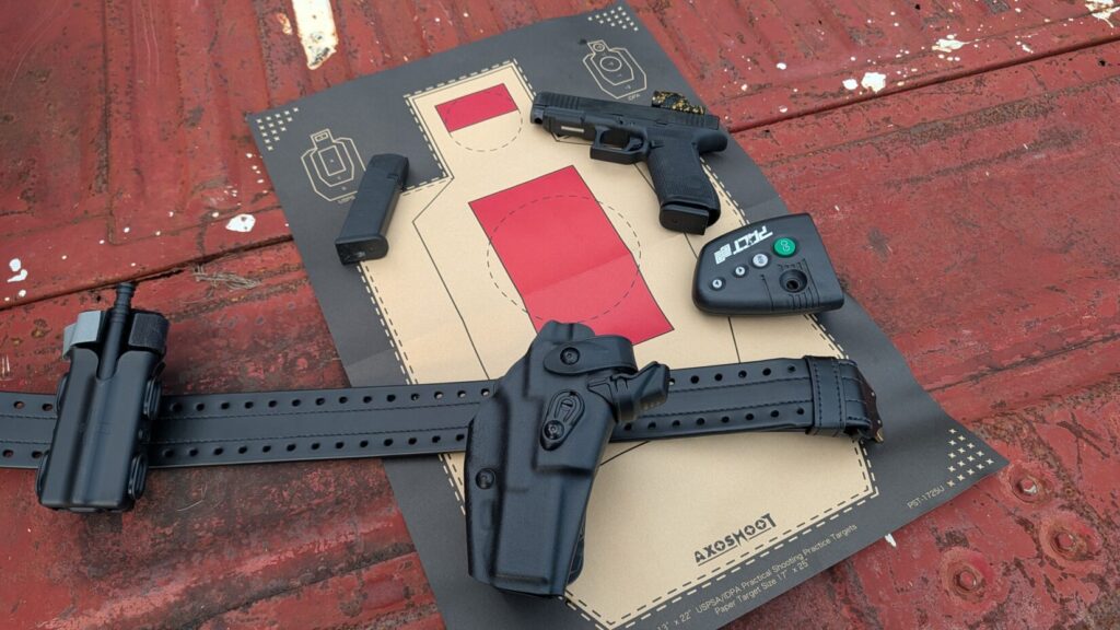 A flat lay on a red surface featuring a paper silhouette shooting target, a black HOLLE belt with a holster and tourniquet, a black handgun with a red dot sight, a spare magazine, and a shot timer.