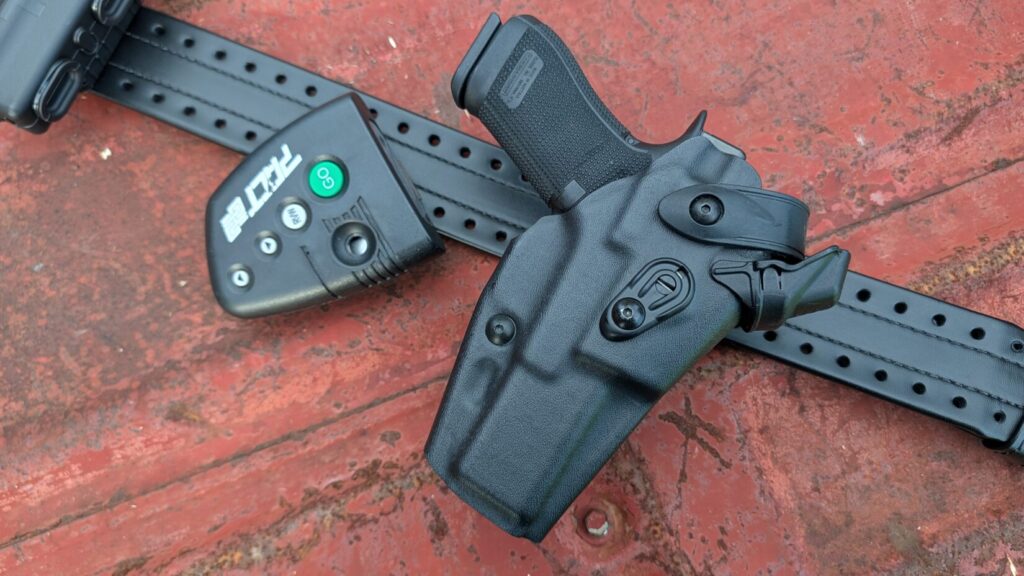 A black pistol secured in a black retention holster attached to a duty belt, placed next to a shot timer on a rusted metal surface.