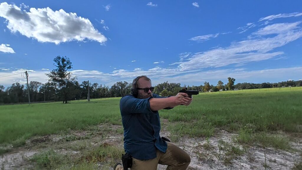 A bearded man kneeling in a grassy field, aiming a handgun with both hands while wearing ear and eye protection.