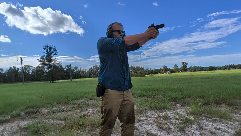 A bearded man standing in a grassy field aiming a handgun with both hands, wearing a holster, ear protection, and sunglasses.
