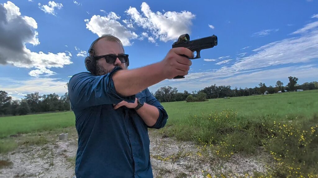 A bearded man wearing safety gear aiming a handgun with one hand outdoors, keeping his other hand pressed against his chest