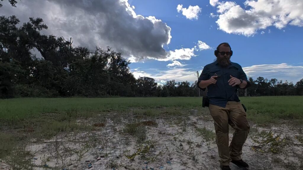 A bearded man wearing safety gear moving quickly across a grassy outdoor shooting range, his hand near his hip.