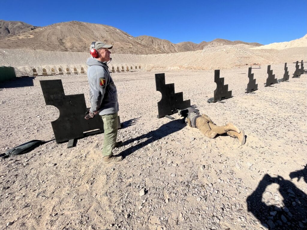 Clint Smith stands on a desert firing range near Las Vegas, Nevada. There is a row of stepped plywood barricades set up about 50 meters from a row of targets. A single student, rolled onto his side, shoots through a gap under a barricade.