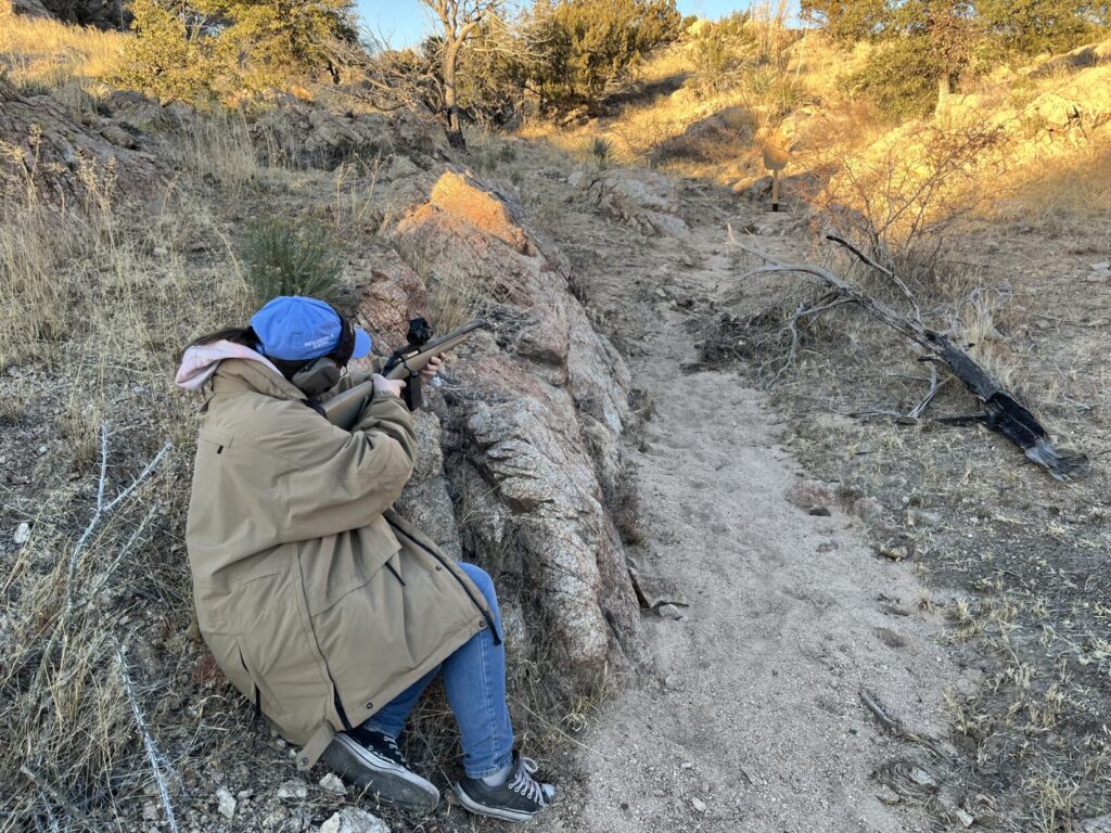 A woman in a blue hat and a tan jacket aims a Ruger American bolt-action rifle from a semi-seated psition behind a rock at a cardboard target about 20 meters farther up an arroyo, or dry watercourse, in hilly, Sonoran Desert. There is a small, zero-magnification optic on her rifle.