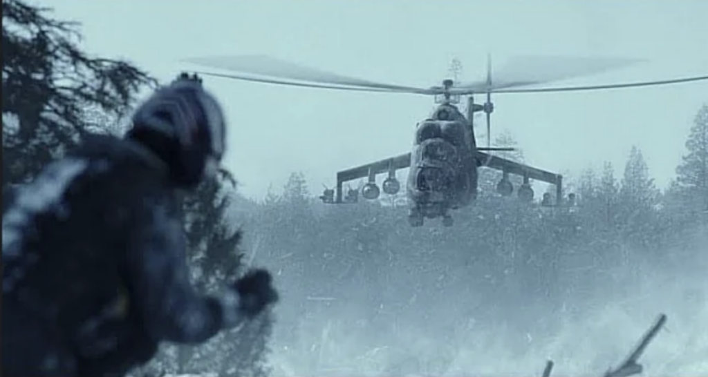 In this image from the motion picture Top Gun Maverick, a US naval aviator in a flight suit and helmet stands in a snowy alpine meadow, on the left side of the screen. A massive "Hind" helicopter gunship is pointed at him. Hinds have short wings called sponsons that project from the fuselage at a slightly downward angle. 