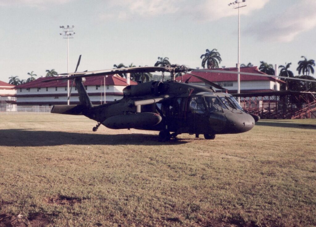 A Blackhawk helicopter sits in a baseball field in front of white buildings with red tile roofs at Albrook Air Force Station in Panama, during Operation Just Cause. Small wings, called sponsons, slant down and out from the top of the fuselage, holding fuel tanks.